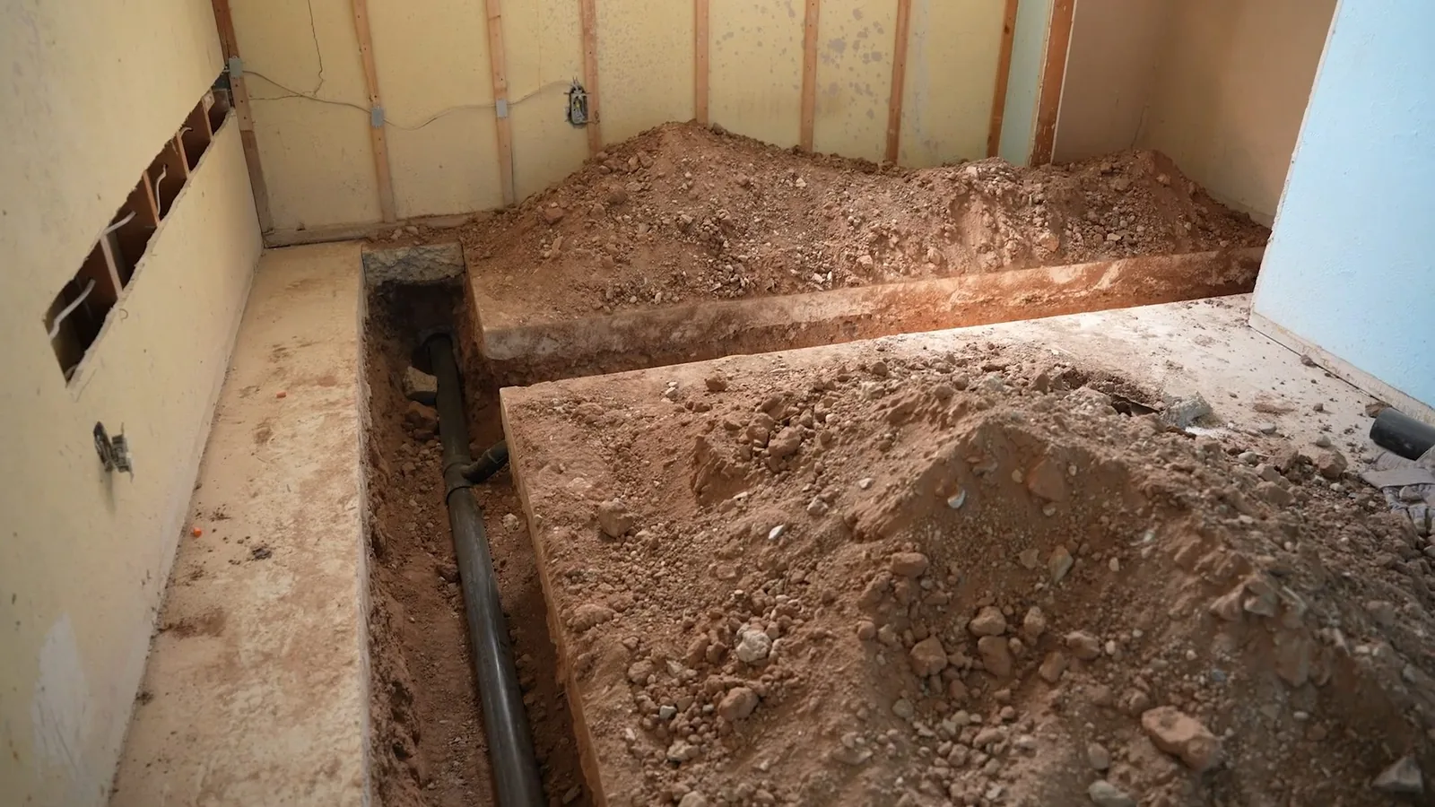 Basement floor opened along the wall with a trench in the soil and dark pipe at the bottom.
