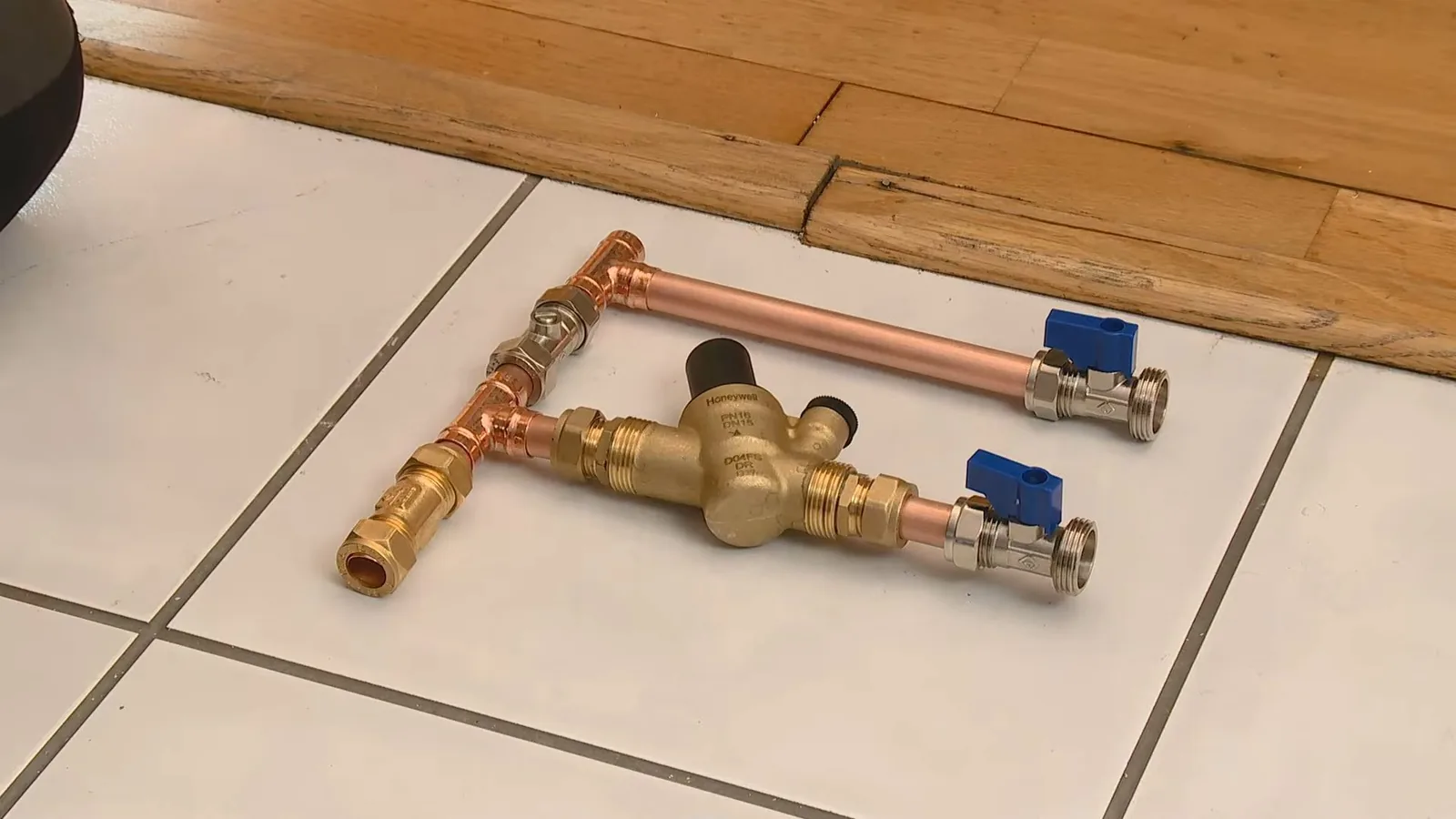 Copper pipe assembly with two blue-handled ball valves and a brass valve between them, laid out on a tile floor.