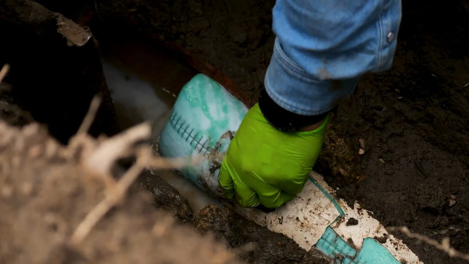 Gloved hand guiding a green liner into an exposed white pipe in a dirt trench.