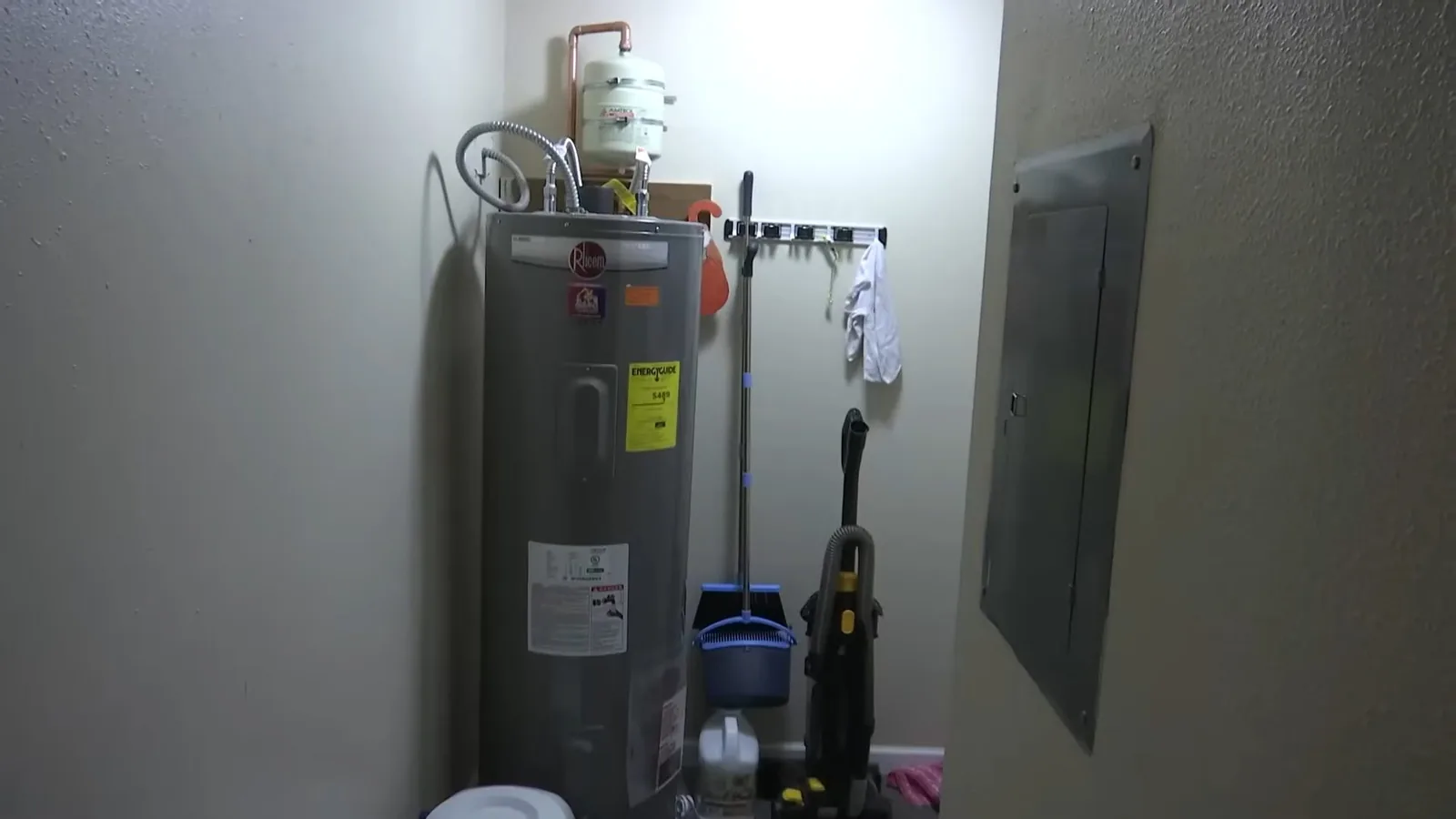 Gray tank water heater with a small white tank on the pipes above it, standing in a utility room beside an electrical panel.