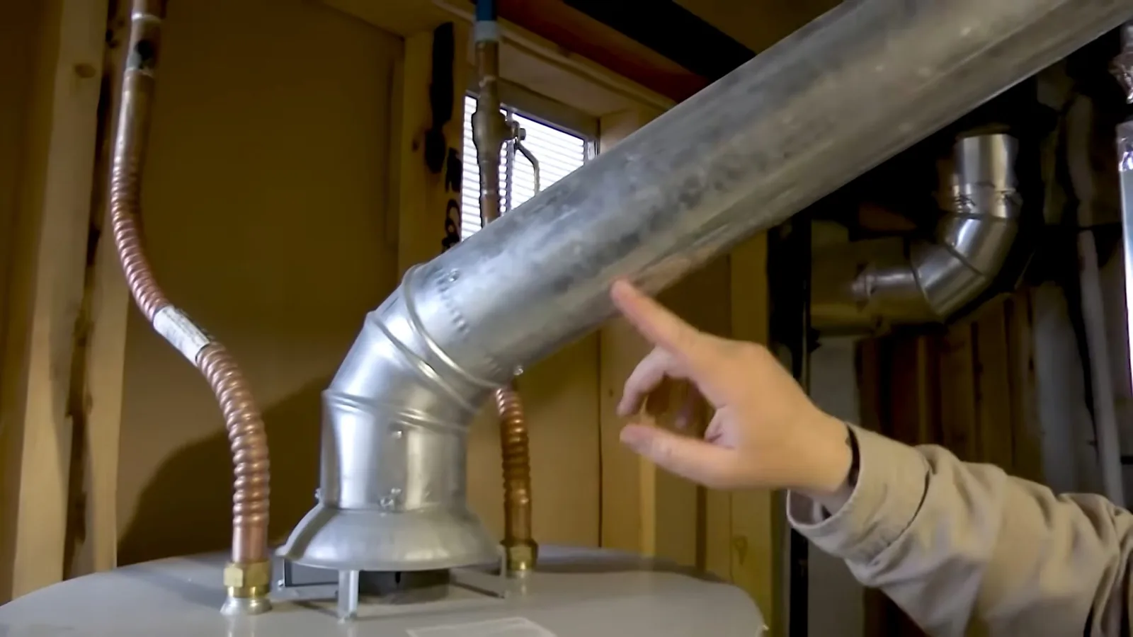 Hand pointing at a galvanized vent pipe rising from the top of a water heater between wall studs.