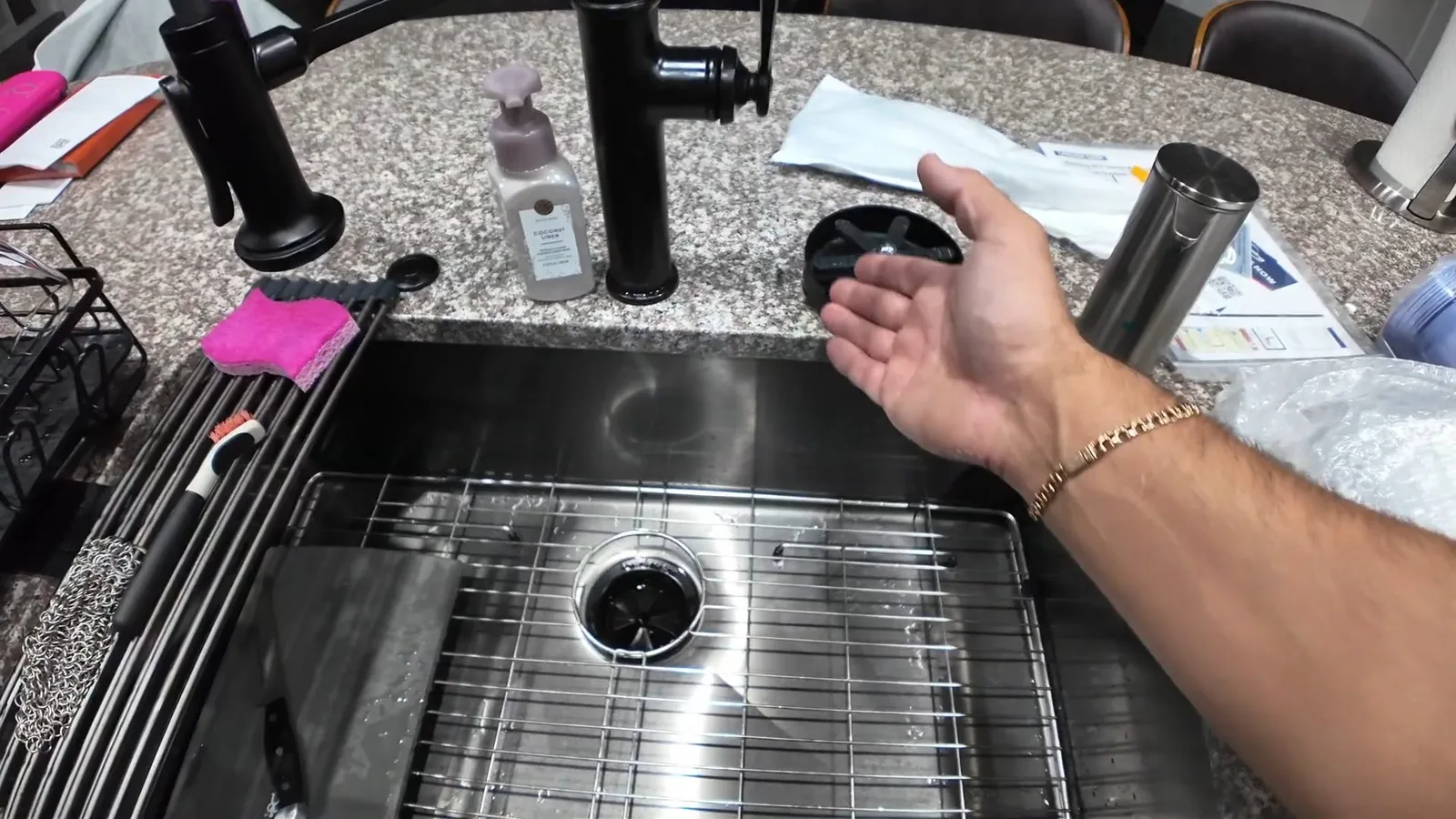 Hand resting beside a steel kitchen sink with a drain strainer, with a black faucet and loose parts on the counter.