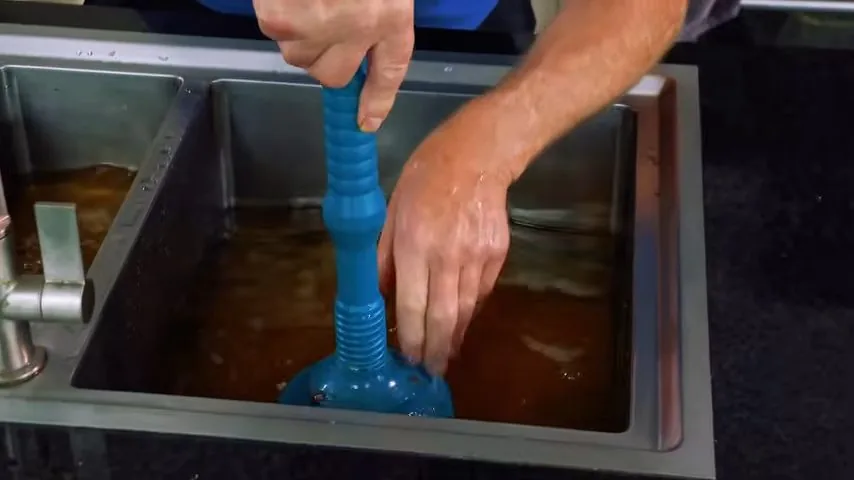 Hand working a blue plunger in a steel sink filled with brown water.