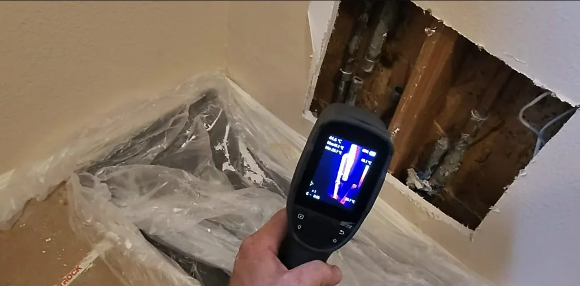 Handheld thermal camera held below a square opening cut in a wall, with plastic sheeting on the floor.