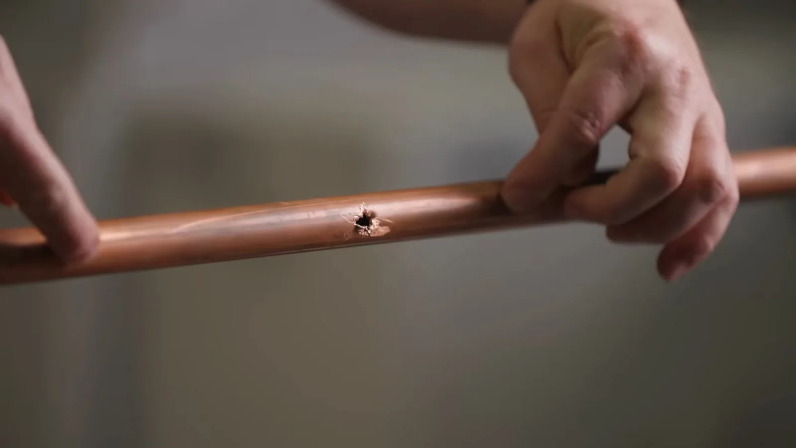Hands holding a length of copper pipe with a small pinhole in its side.