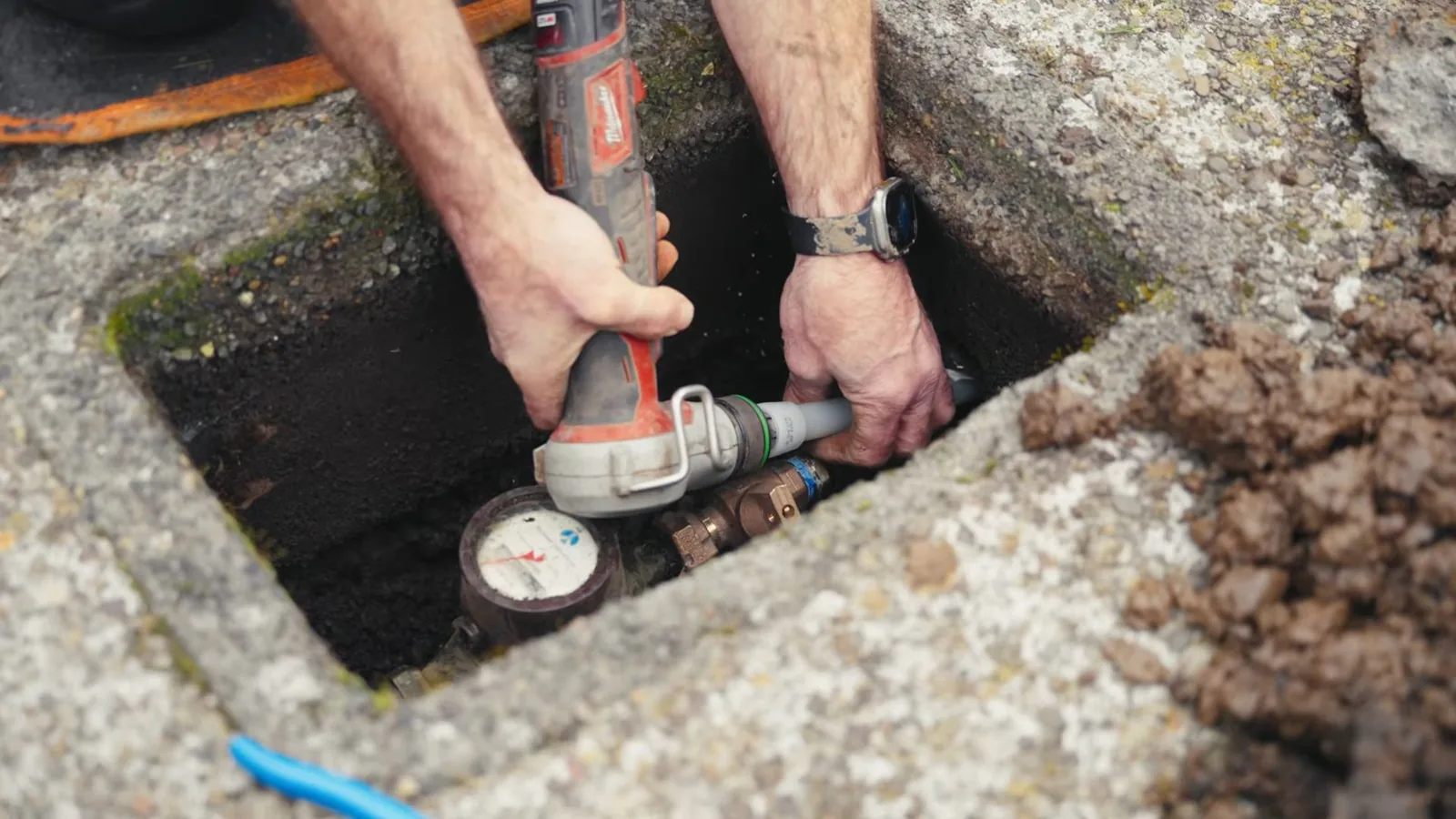 Hands using a power tool on a pipe next to a water meter inside a ground-level box.