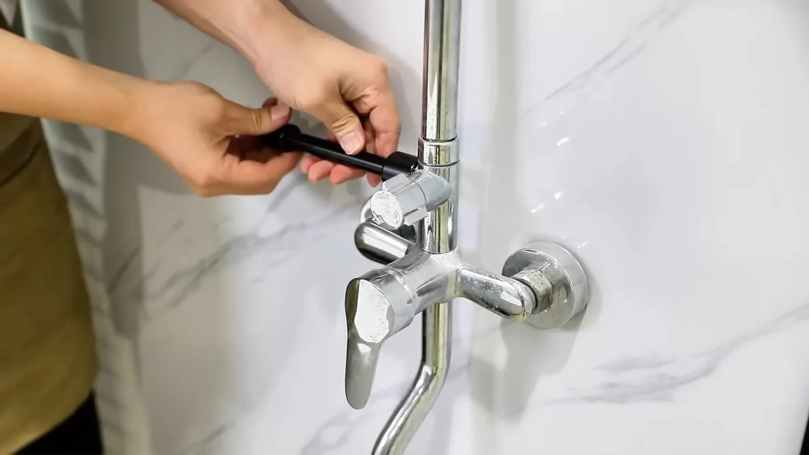 Hands working on a chrome wall-mounted faucet and shower riser on a marble-look wall.