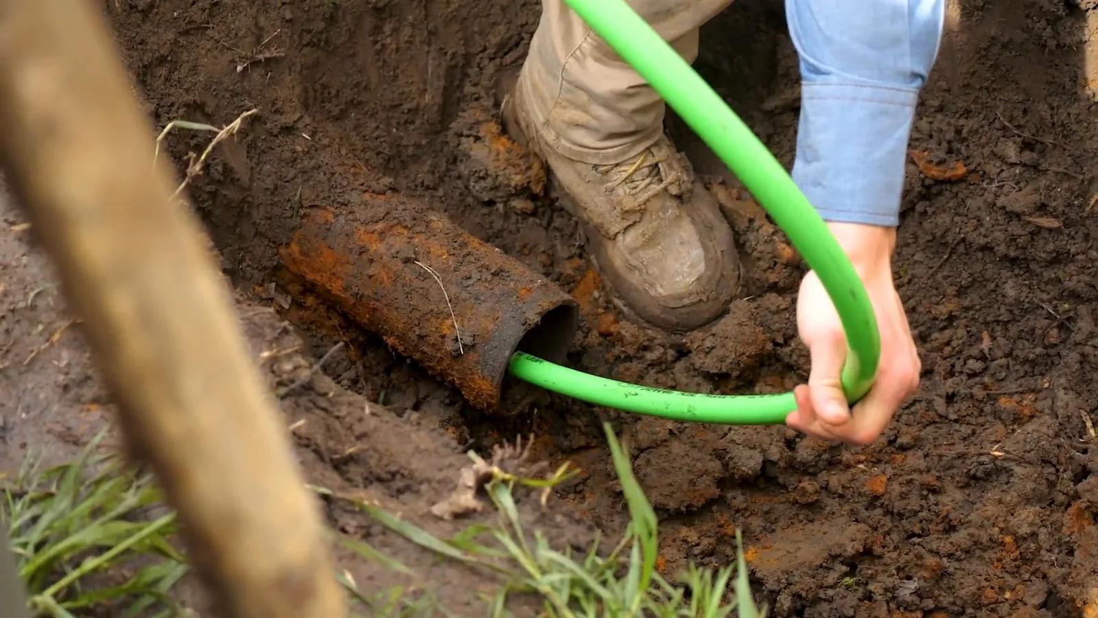 Gloved hand guiding a green liner tube into an exposed rusted sewer pipe in a trench.