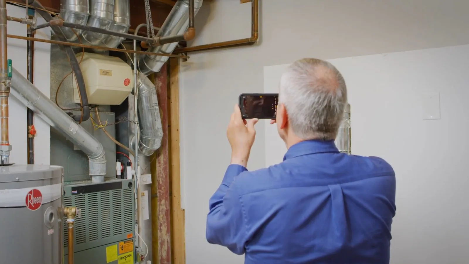 Man in a blue shirt photographing a tank water heater and furnace with his phone.