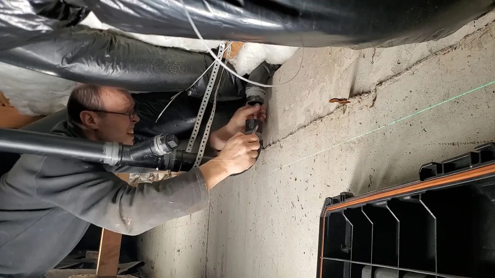 Man in a crawlspace looking at a pipe under black-wrapped ductwork, with a green laser line across the wall.