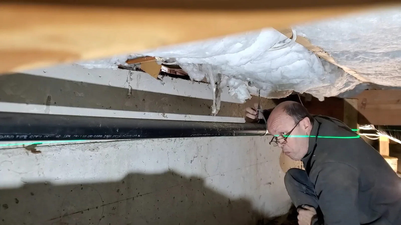 Man crouching in a crawlspace beside a black pipe, with torn insulation hanging from the floor above him.