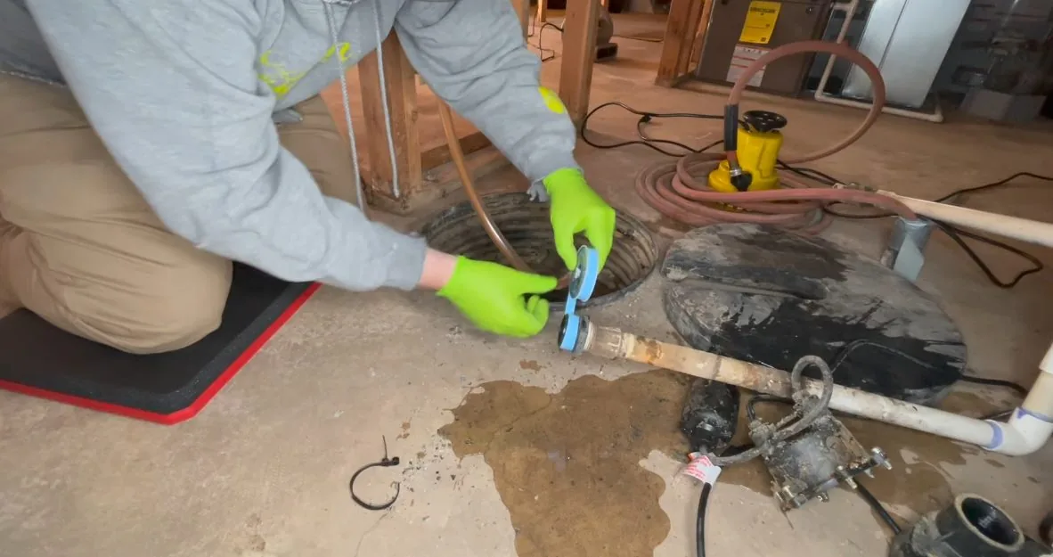 Man in green gloves working on a sump pump pipe in a basin in a basement floor, with a stain on the concrete.