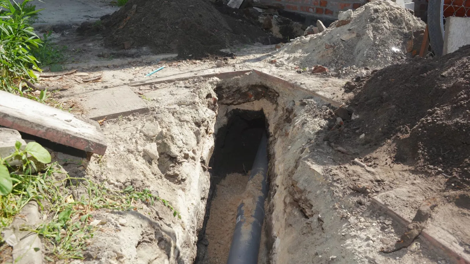 Narrow trench dug beside a building with a dark pipe running along the bottom.