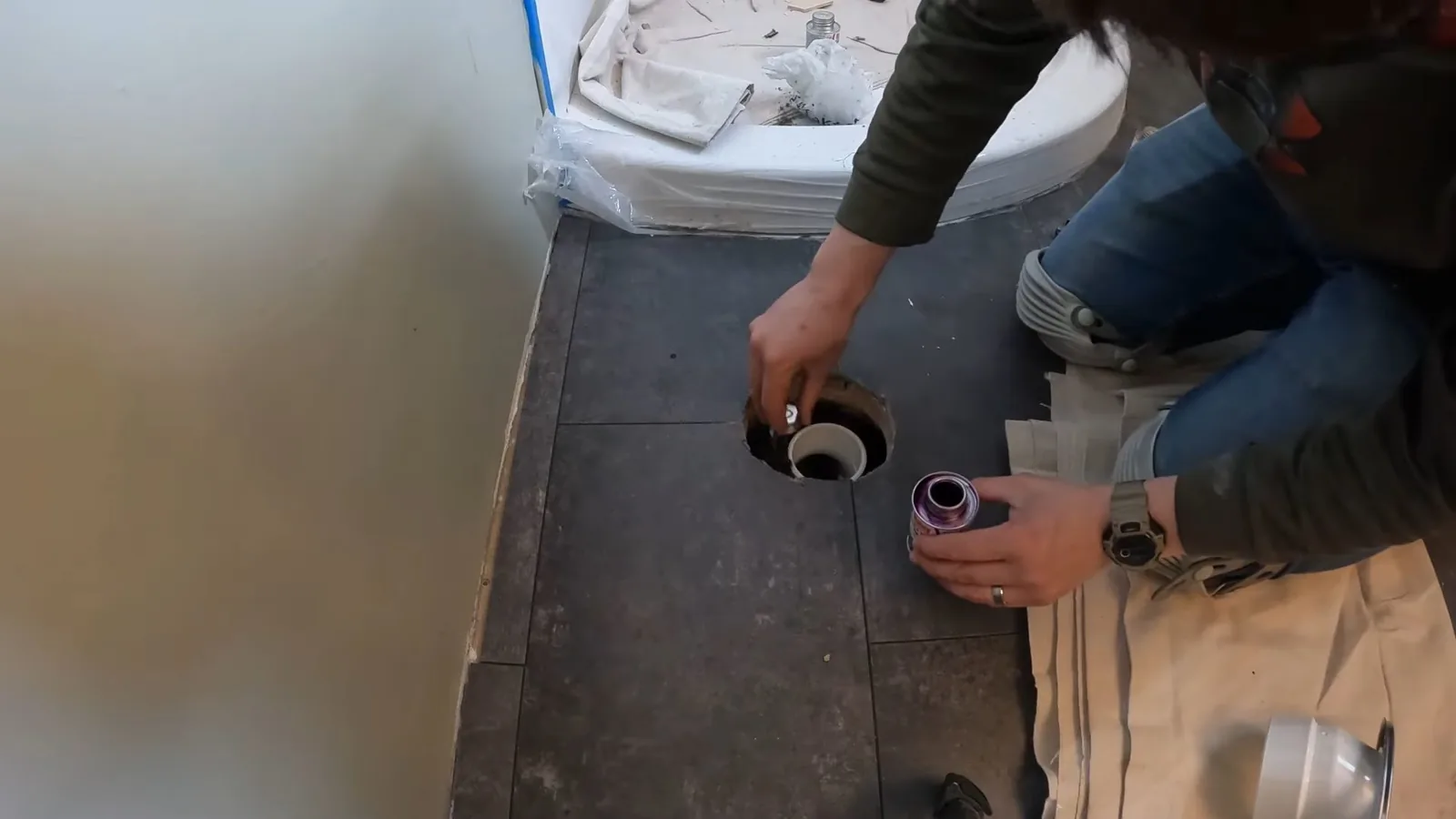 Person kneeling beside a round drain opening in a bathroom floor with the toilet removed.