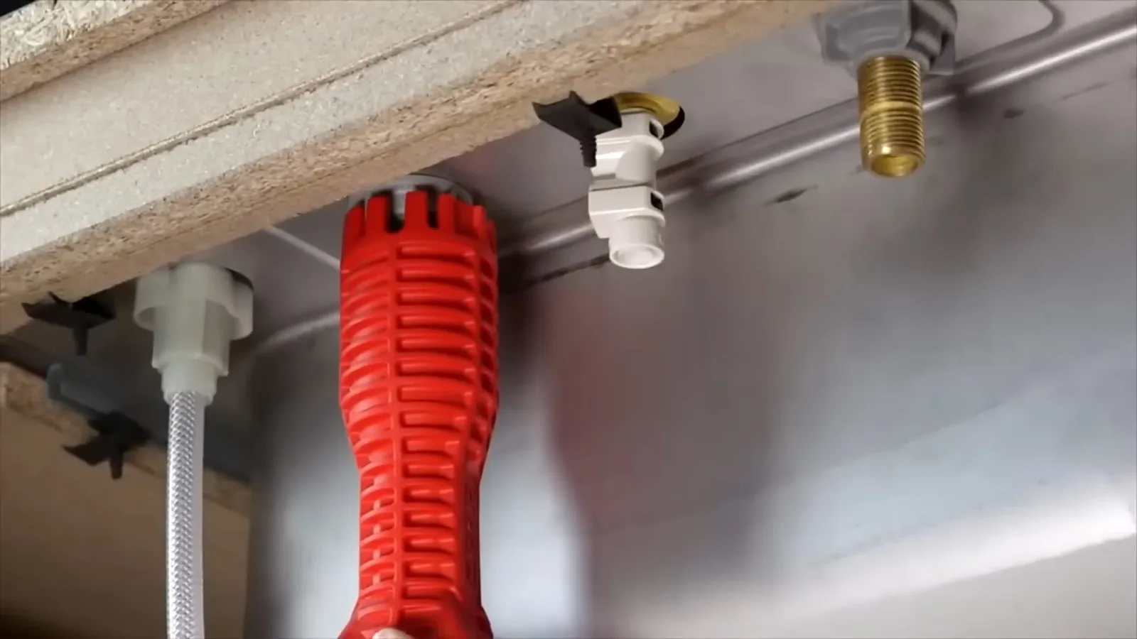Red plastic wrench held under a stainless sink near a white drain hose and a supply fitting.