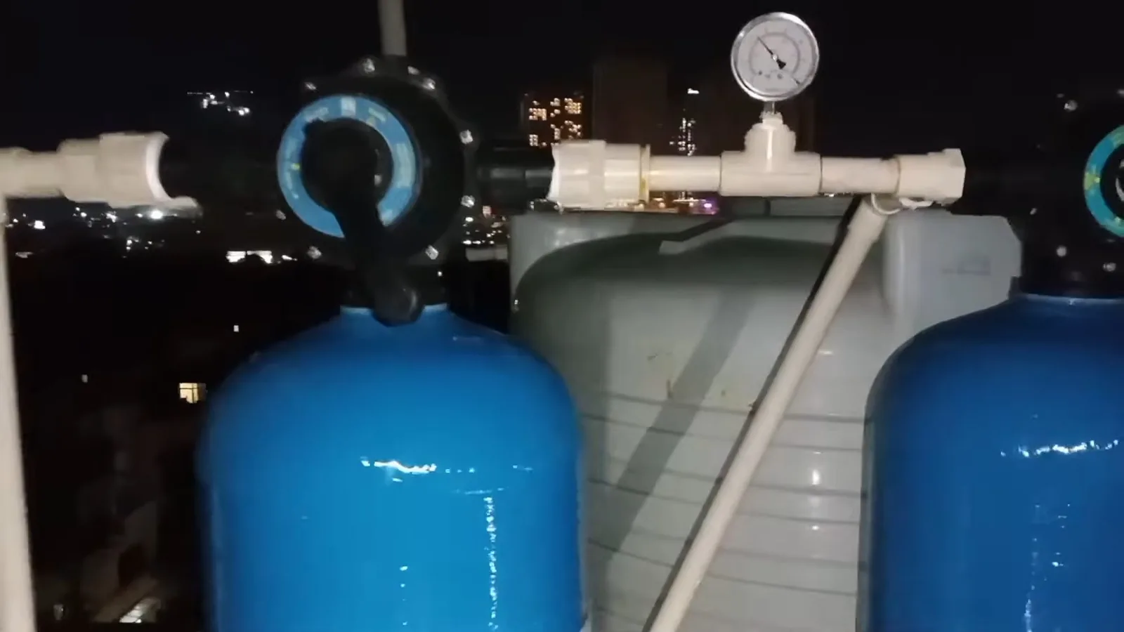Two blue pressure tanks with a gauge on the white pipe between them, with city lights in the background.