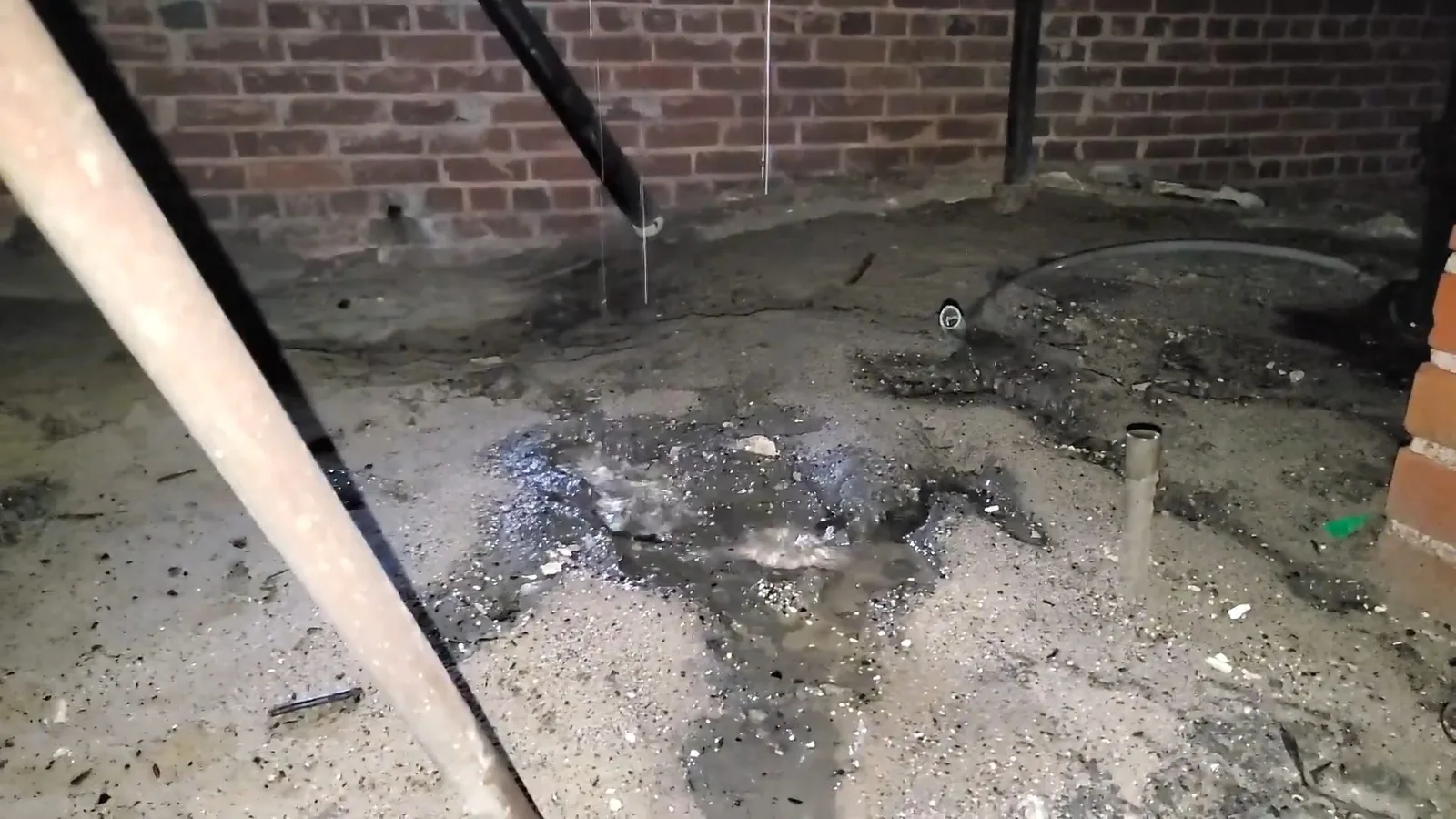 Wet gravelly ground under a crawlspace, with a brick wall behind it and a pipe running diagonally through the foreground.