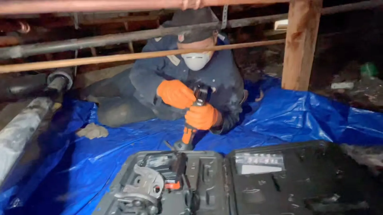 Worker in a mask and orange gloves kneeling on a blue tarp in a crawlspace under pipes, with a tool case beside them.